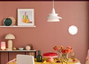 Modern dining room with two‑tone pink and plum walls and a white pendant light. A wall shelf holds framed art and ceramics, a dining table and chairs with a mustard tablecloth, glass vases and a red cake stand.
