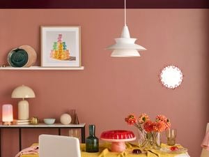 Modern dining room with two‑tone pink and plum walls and a white pendant light. A wall shelf holds framed art and ceramics, a dining table and chairs with a mustard tablecloth, glass vases and a red cake stand.