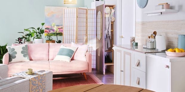 A styled open-plan living and kitchen space featuring a pink sofa, folding room divider, indoor plants and light pastel furnishings.