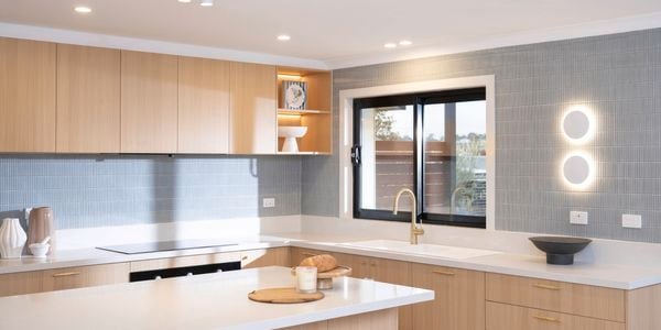 A modern kitchen features light wood cabinetry, a sleek white benchtop, vertical tile splashback, and stylish lighting.