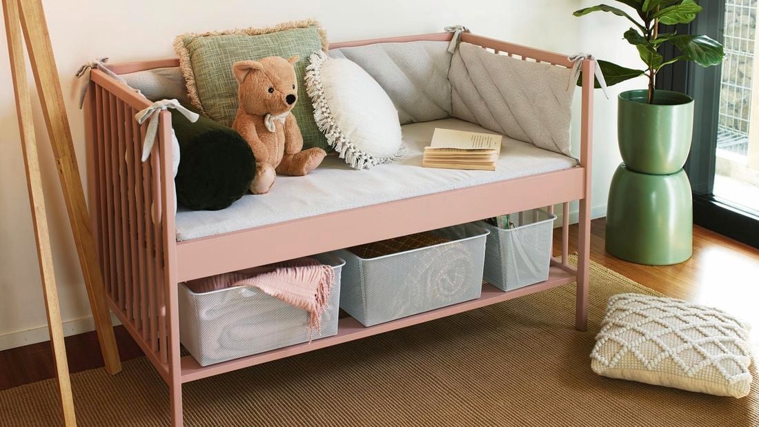 Completed light pink reading nook carrying grey pillows and an open book. 2 Grey mesh storage containers carry a blanket and books underneath the seats
