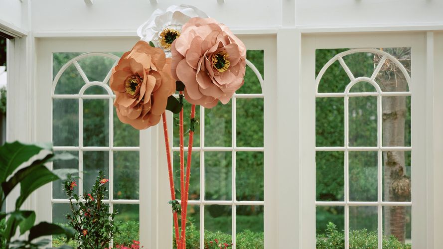 Three giant paper flowers in peach and white colours, surrounded by potted plants in a sunlit room with large windows.