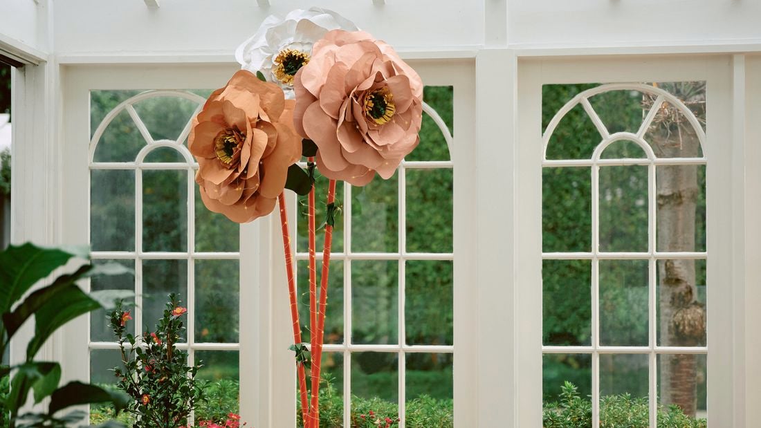 Three giant paper flowers in peach and white colours, surrounded by potted plants in a sunlit room with large windows.