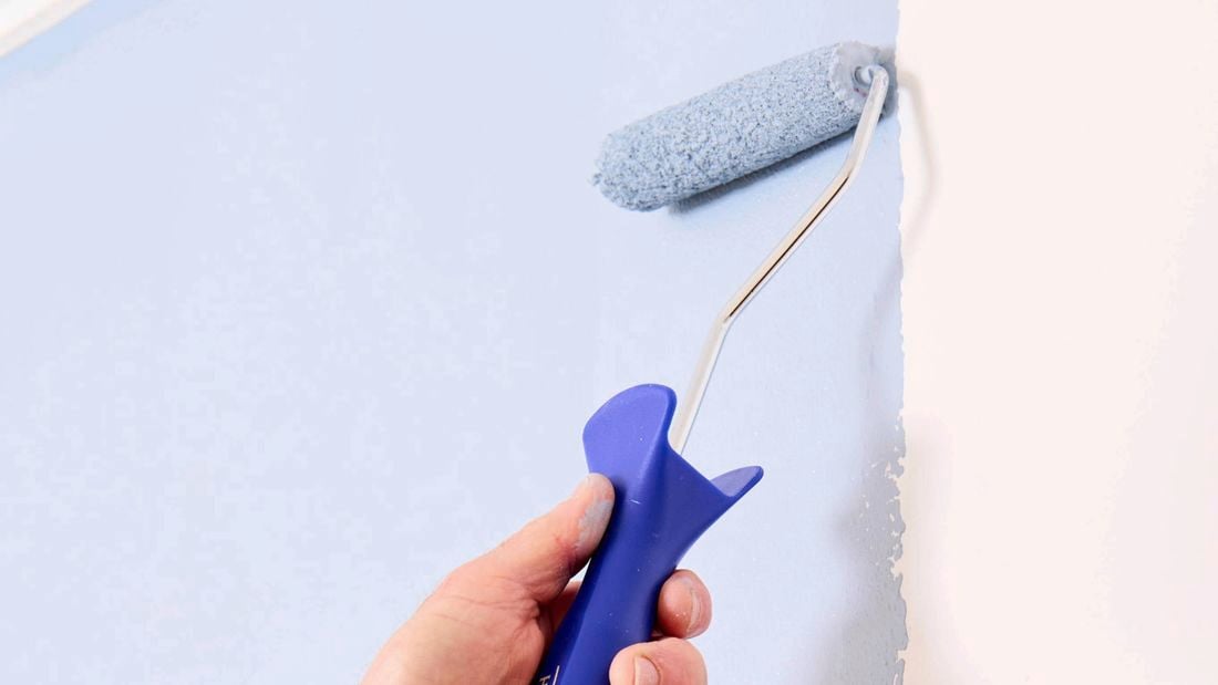 A hand holding a blue Monarch mini paint roller, painting pale blue paint onto a white wall.