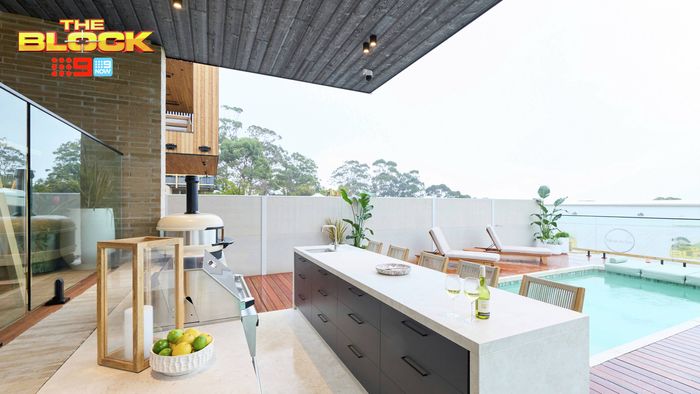 Covered outdoor kitchen and pool area featuring a large island bench, barbecue, timber decking and poolside seating.
