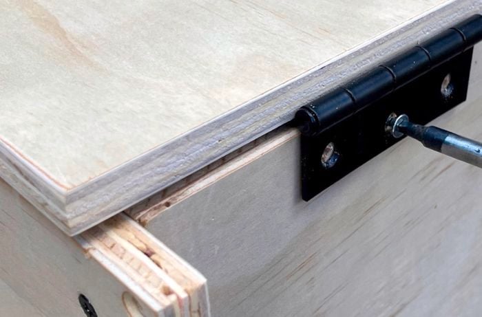 A wooden box with a hinge being attached to the side of a panel with a screwdriver