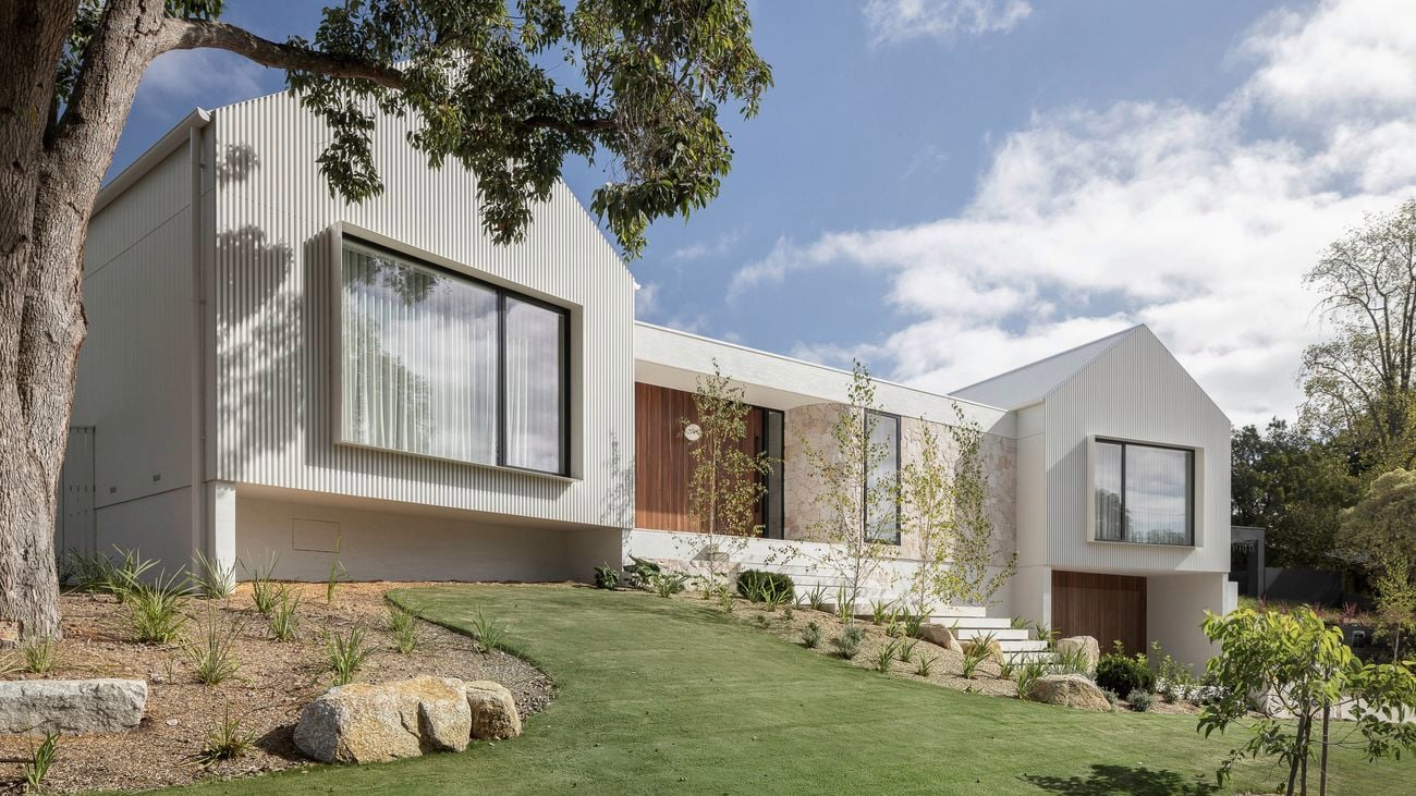 Modern architectural home with light vertical cladding, stone and timber accents, large windows, and a sloped landscaped garden with trees and rock features under a blue sky.