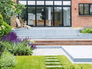Modern backyard garden with patio, steps, plants, and outdoor furniture near a brick house with large windows."