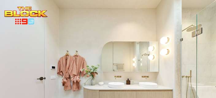 Modern bathroom with a floating timber double vanity, white basins, rounded mirrors, statement wall lights, glass-enclosed shower and pink robes hanging on the wall. “The Block” logo appears in the upper-left corner.