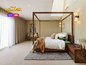 A stylish bedroom featuring a central four-poster bed with white and green bedding, multiple pillows, and a brown throw blanket. A wooden nightstand with books and decor sits to the right, while a cushioned bench with decorative pillows lines the left wall. The room includes a textured rug, light beige walls, wall-mounted lights, and artwork above the bench and bed. Sheer curtains cover large windows, allowing natural light in. 