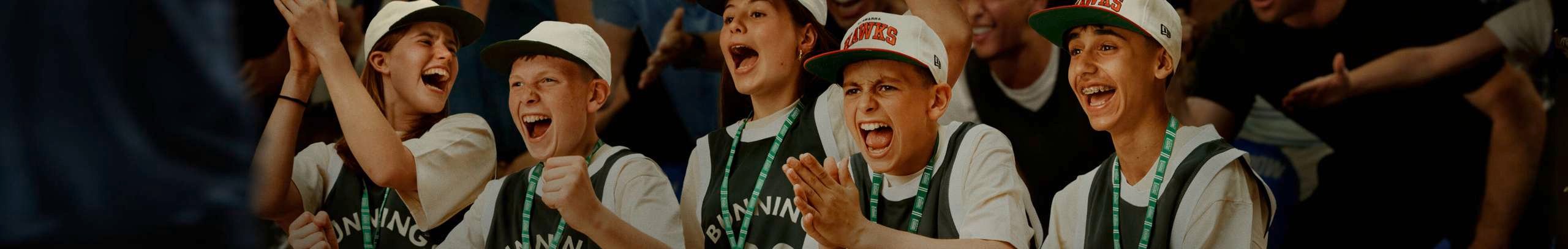 Fans wearing matching jerseys and hats sit on a bench in a crowded basketball arena, clapping and cheering as a player runs past in the foreground.