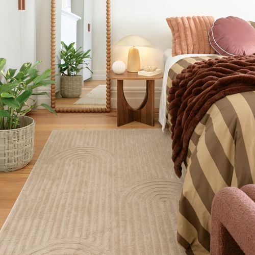 A styled bedroom corner with a bed dressed in warm-toned textiles, a wooden bedside table with a lamp, a beaded-frame floor mirror, and potted plants on timber flooring.
