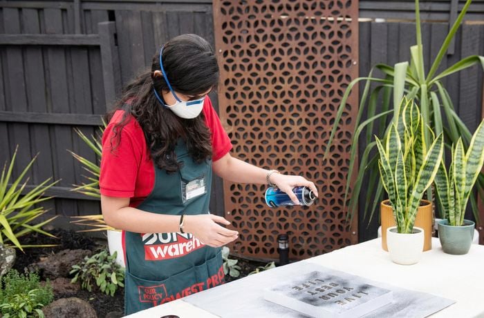 Woman wearing a mask spray painting letters arranged on a stretched canvas