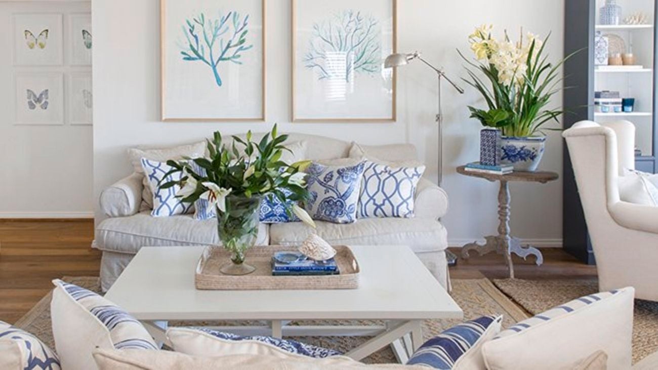 A Hamptons style living area with white couches, blue and white cushions, a white coffee table, wooden floorboards and flowers.