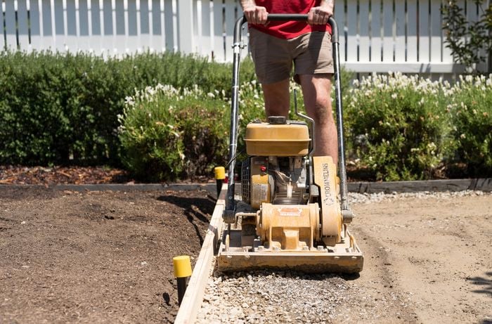 A person in a red shirt and shorts is using a compactor to compact the road base within a framed area. 