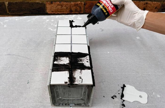 Applying black grout to the joints of a white tiled vase