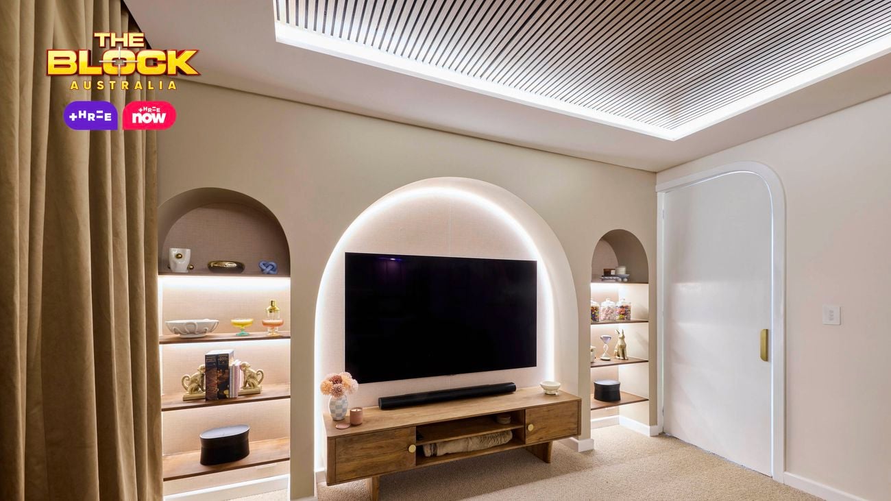 A modern living room with a large flat-screen TV mounted within a softly lit arched alcove. Below the TV is a wooden console table with decorative items and storage compartments. Built-in shelves in arched niches on either side of the TV display vases, books, and sculptures. The ceiling features recessed lighting and slatted paneling for a stylish finish.