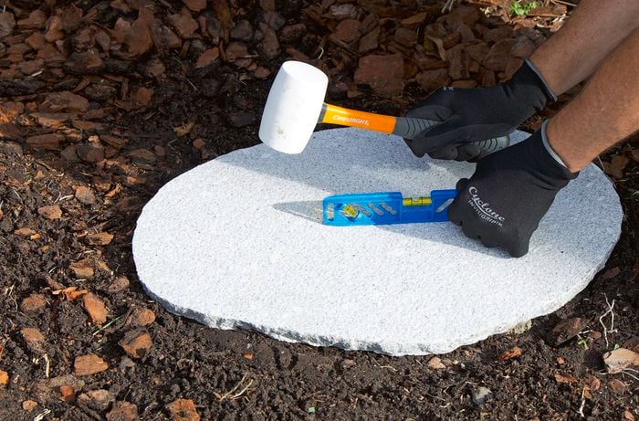 A person wearing gloves uses a rubber mallet and a level on a circular concrete slab in a garden.   