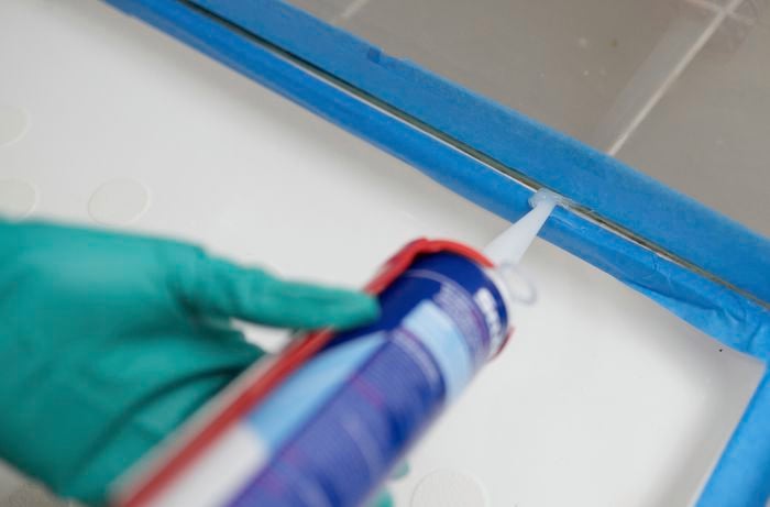 A hand wearing green gloves is applying fresh silicone between the masking tape.