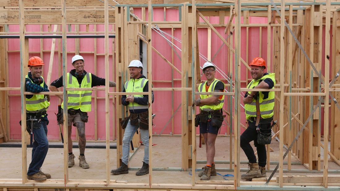 An image of five tradies wearing yellow hi-vis - two in orange hard hats and three in white hard hats - looking to camera through timber frames on a construction site
