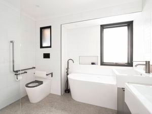 A white tiled bathroom with a bath under a framed window with handrails and toilet with handrails, a sink and a free standing shower