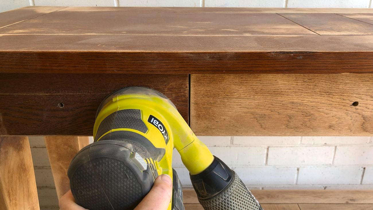 A person using a Ryobi sander to prepare a timber furniture piece for paint.