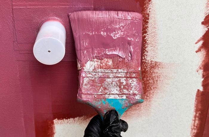 Close up of a black‑gloved hand using a paintbrush to apply pink‑red sealer to a white panel.