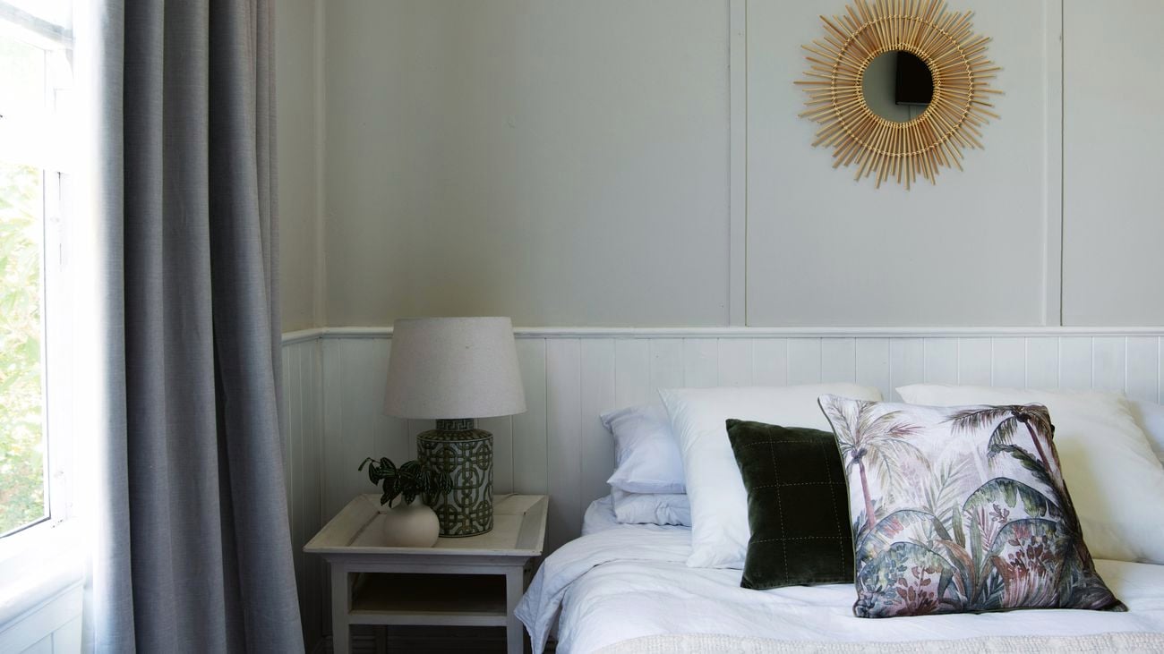A coastal-look bedroom with feature mirror hanging above a bed with no headboard. The bed has a striking palm print cushion displayed amongst other neutral cushions.