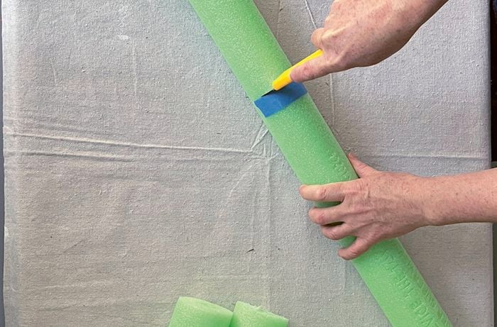 Hands cutting a green pool noodle with a utility knife.