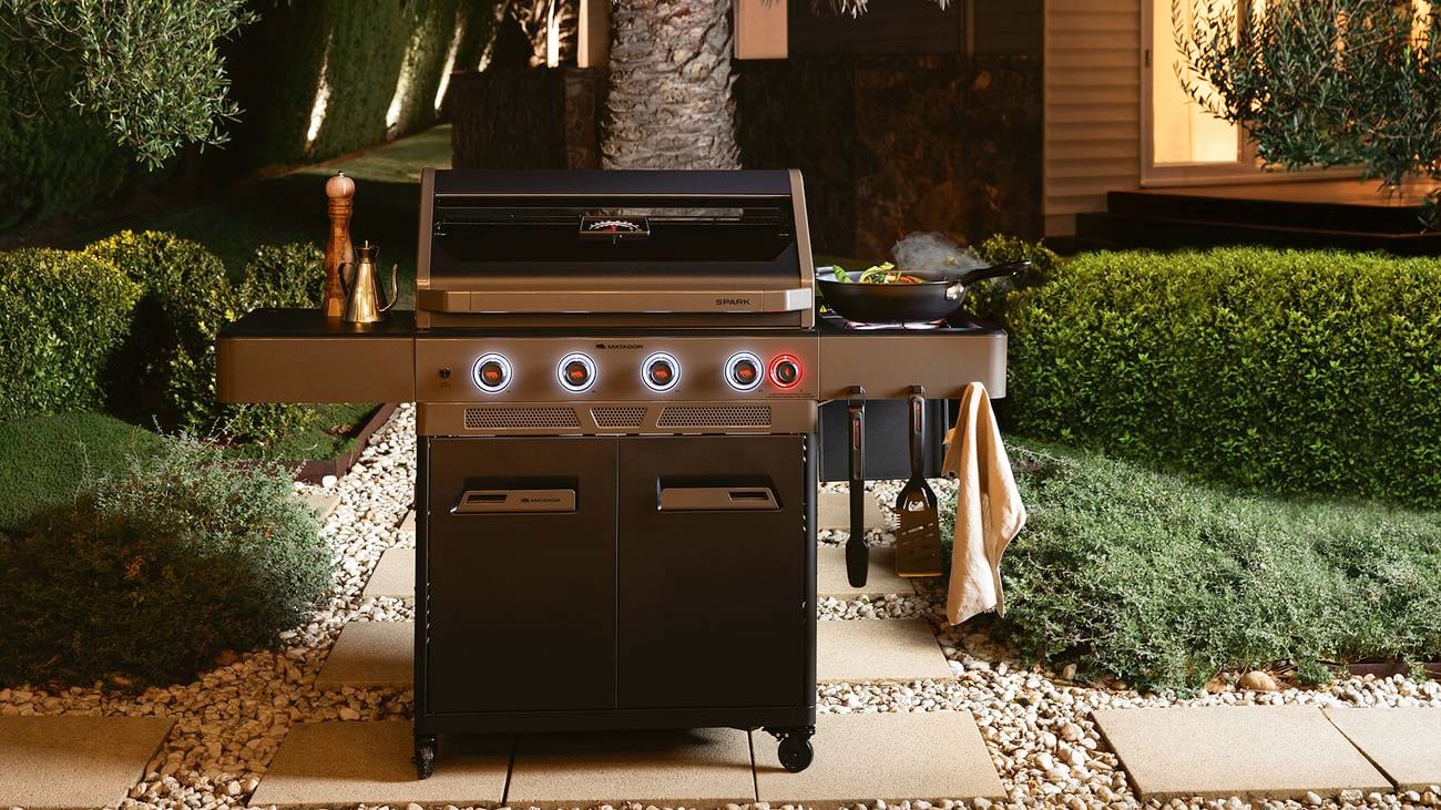 A Matador ‘Spark’ 4 burner gas barbecue with prepared food on the side and condiments sits on some stone paving in a garden at night time.