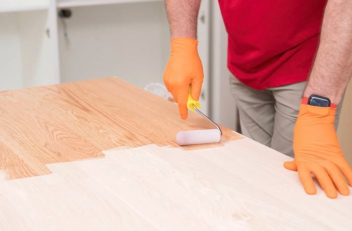 A person wearing orange gloves is applying a first coat of sealant to a wooden benchtop using a paint roller. 