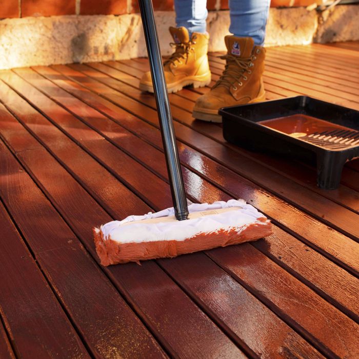 Applying timber deck stain with a deck applicator pad on a wooden outdoor deck. A paint tray sits nearby while a person wearing work boots stands in the background, showing the process of staining and protecting outdoor timber decking.