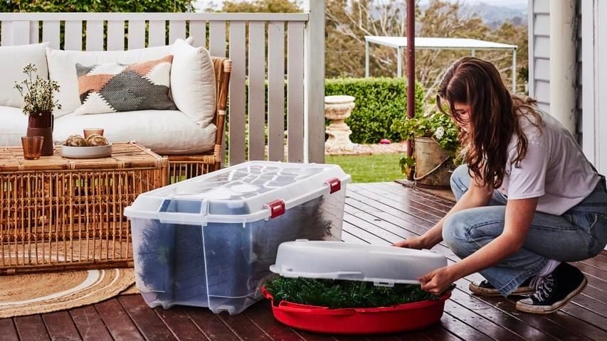 Person crouching on a verandah, closing the lid to a large round plastic container with a christmas wreath inside.