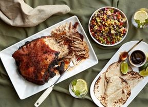 Smoked pulled pork on a platter with a serving fork, a bowl of cowboy caviar and a plate of tortillas with lime wedges and barbecue sauce