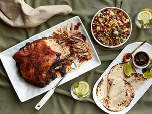 Smoked pulled pork on a platter with a serving fork, a bowl of cowboy caviar and a plate of tortillas with lime wedges and barbecue sauce
