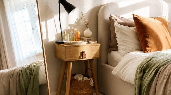 Timber bedside table beside an upholstered bed, styled with a lamp, decor accessories and layered neutral bedding.
