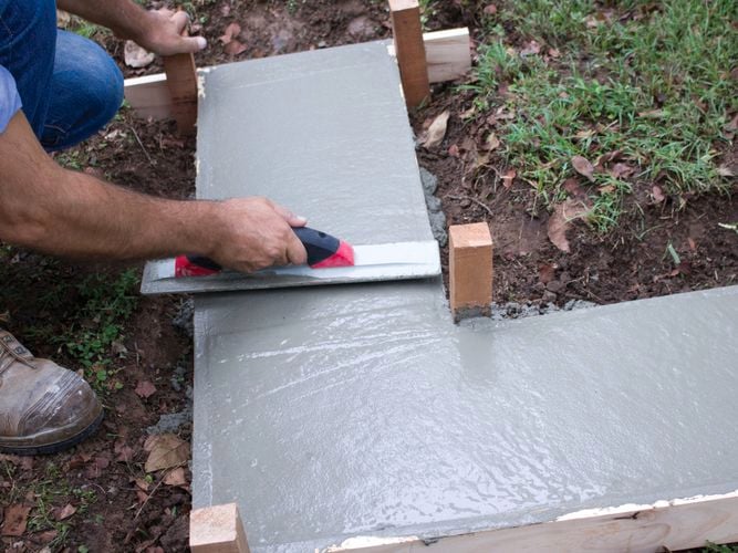 Person smoothing freshly poured concrete with a trowel inside a wooden frame, surrounded by grass and soil.
