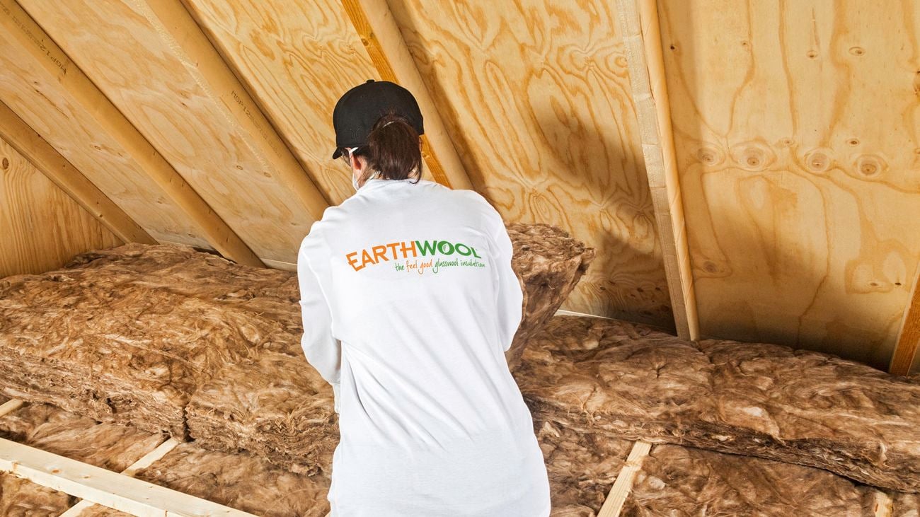 A person kneeling in the wooden structure of a ceiling, facing away while installing a batt of Earthwool insulation in the ceiling