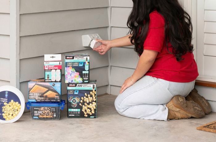 A woman in a red t-shirt is plugging a double adaptor into a weatherproof power point, while some lights are lying on the floor.