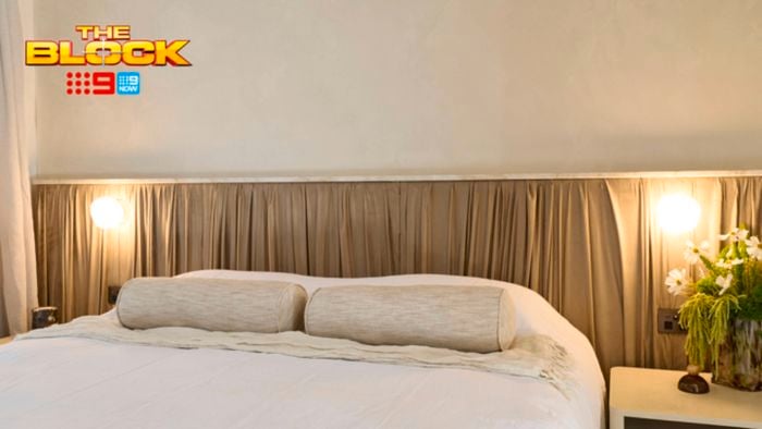A furnished bedroom with a bed, side table with a vase on it and a bedhead behind it. And The Block logo in the image.
