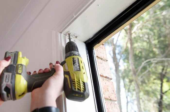 A person attaching a bracket at the top of a window opening using a cordless drill