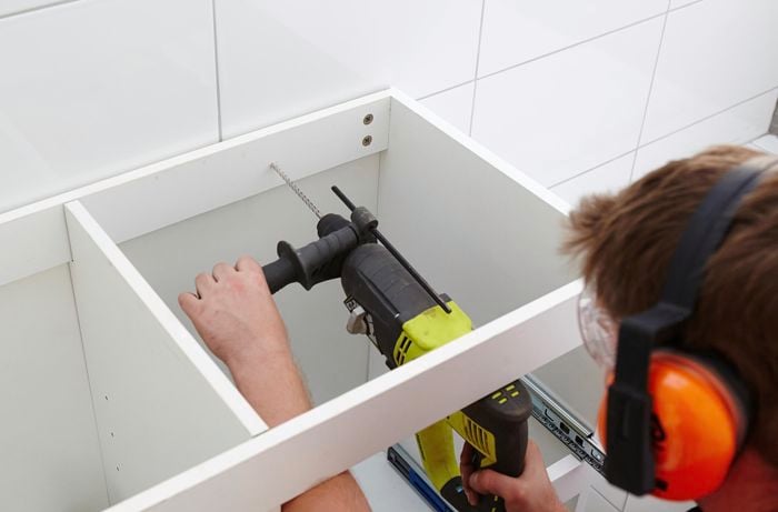A person using a hammer drill to drill a hole through the back rail of a cabinet into a wall 