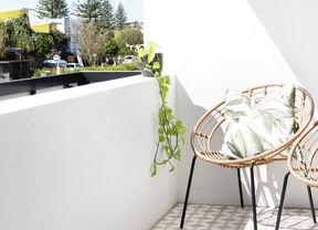 A bright balcony with patterned floor tiles, two wicker chairs, and a view of trees and buildings under a clear blue sky.