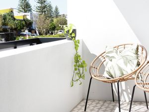 A bright balcony with patterned floor tiles, two wicker chairs, and a view of trees and buildings under a clear blue sky.