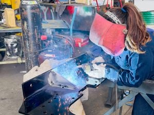 A person wearing protective welding gear, including a large face shield and gloves, is welding a black metal component on a workbench. Bright sparks and blue light from the welding torch illuminate the workspace. The surrounding area contains gas cylinders, tools, and industrial equipment, indicating a workshop environment.