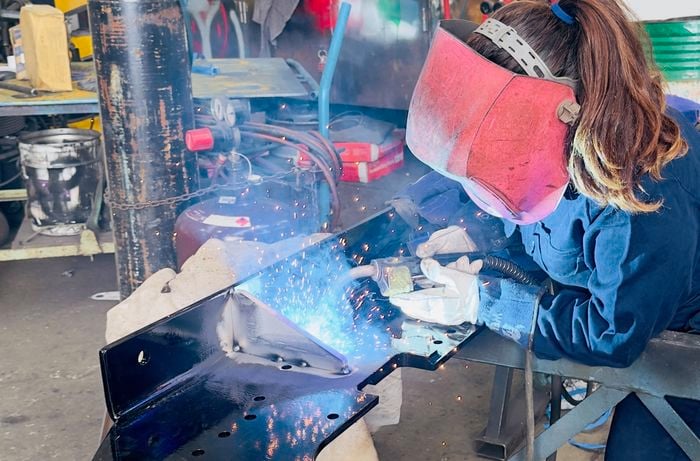 A person wearing protective welding gear, including a large face shield and gloves, is welding a black metal component on a workbench. Bright sparks and blue light from the welding torch illuminate the workspace. The surrounding area contains gas cylinders, tools, and industrial equipment, indicating a workshop environment.