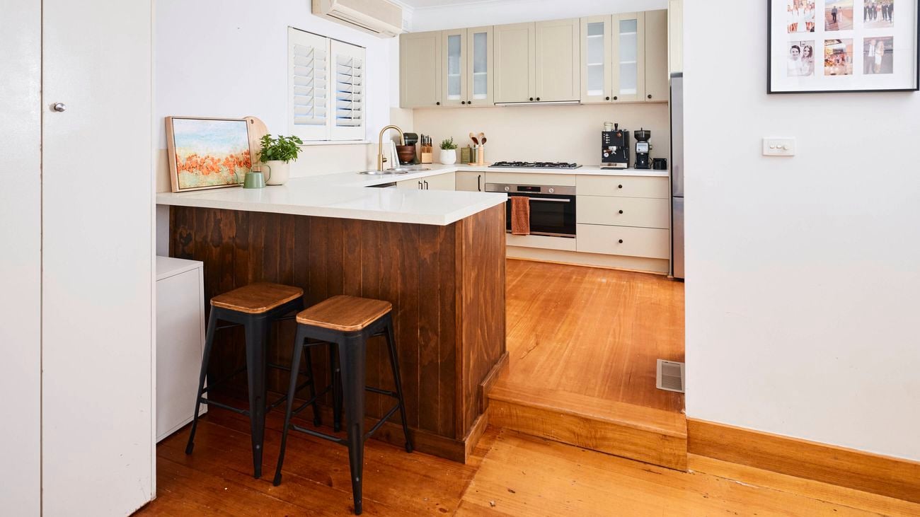 Open-plan kitchen with a white island bench and dark wood base, two bar stools, beige cabinetry, and a cooking area in the background, featuring warm timber flooring and natural light from a skylight.