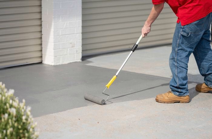 A person in red shirt and work boots is applying the paint on the concrete floor using a paint roller.