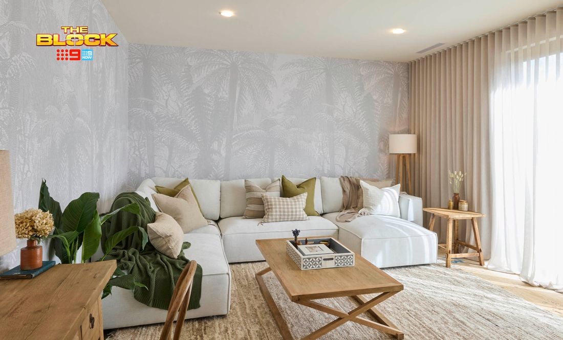 A modern living room with a large white sectional sofa decorated with neutral and green pillows. A wooden coffee table with a decorative tray sits in front, and a side table with a lamp and vase is placed to the right. Light-colored curtains cover large windows, and palm tree wallpaper adds texture to the walls. A desk with drawers and a chair is positioned on the left, near potted plants. 