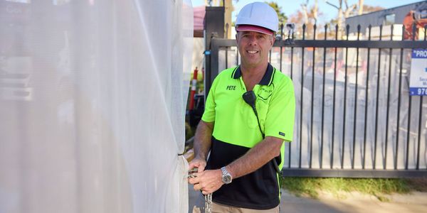 A man in a yellow hi-vis t-shirt and white hardhat looks at the camera while holding a lock chain beside a fence on a construction site